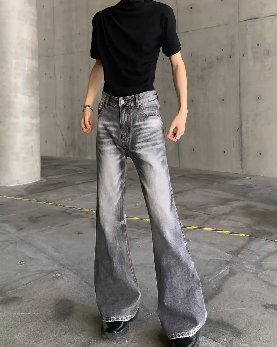メンズのグレーのウォッシュ加工フレアデニムパンツ。裾の擦り切れと膝下の広がりを写した、柱を背にした正面の着用写真。