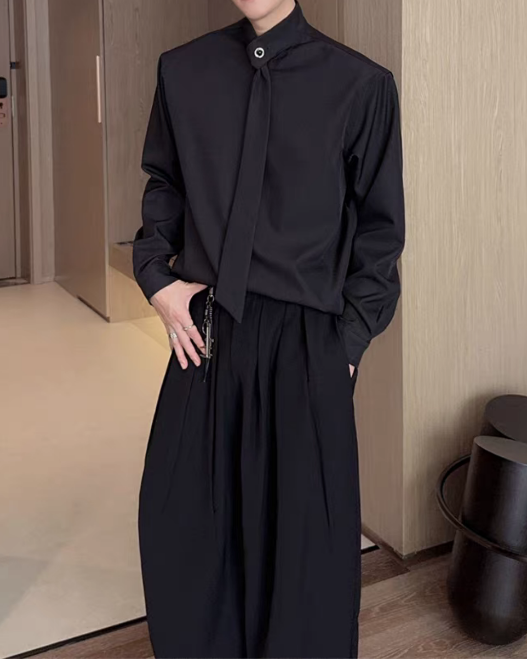 黒のシャツ。スタンドカラーにタイデザインの装飾が付き、長袖。ワイドなドレープを持つパンツとセットアップで、モノトーン配色。