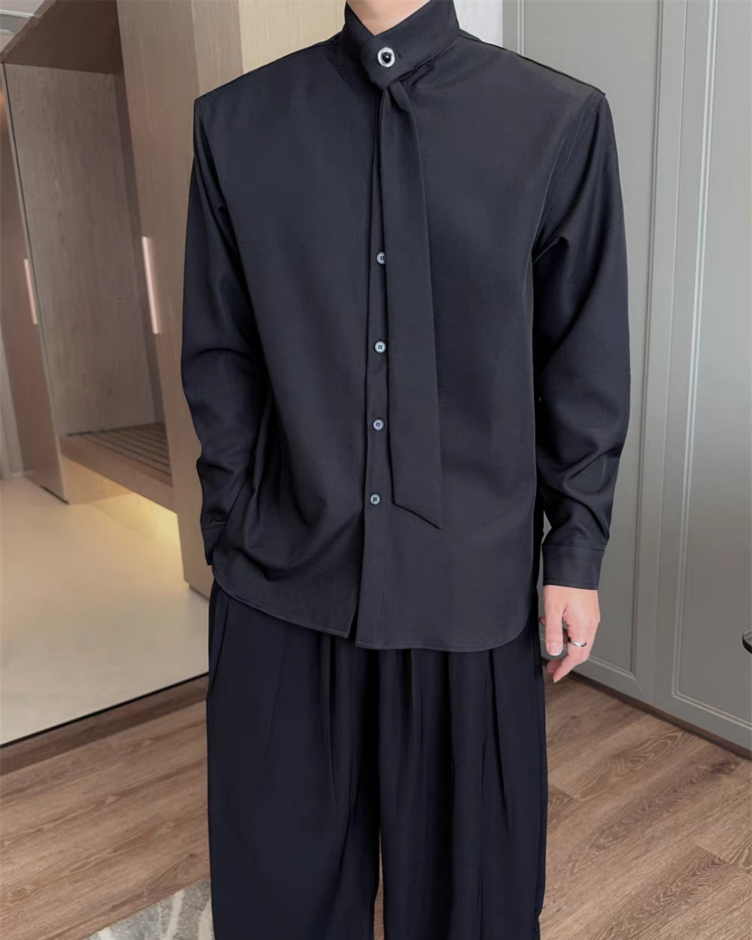 黒いシャツに結びモチーフの装飾タイが付き、スタンドカラーと白いボタン留めの仕様。長袖のオーバーサイズシルエット。