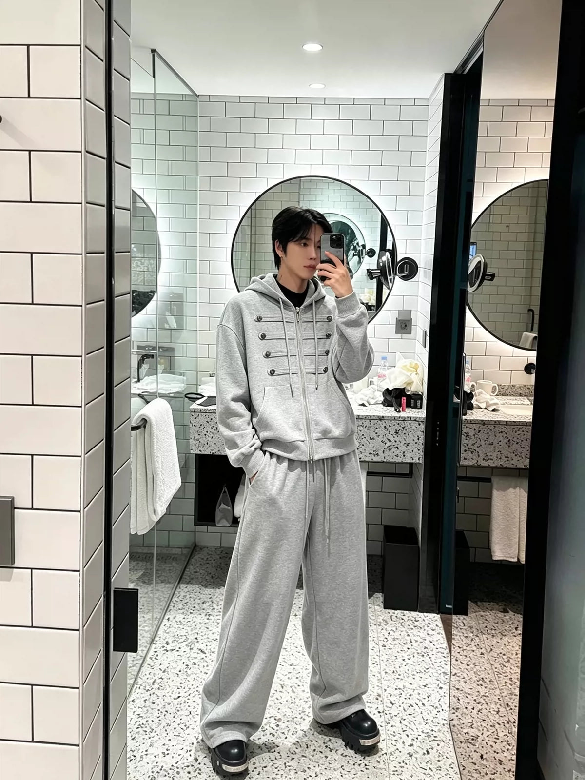 メンズのグレーのセットアップを鏡越しに写した全身。短め丈のジップパーカーと、足元まで落ちるワイドなスウェットパンツ。