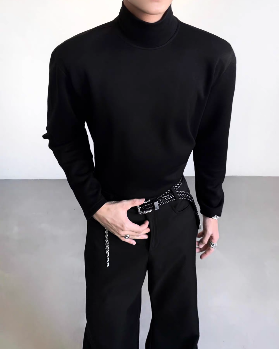 メンズの黒のタートルネックカットソーを正面から寄りで写した着用写真。高さのある襟と肩の張り、細身の長袖が分かり、下は黒パンツと編み込みベルト。