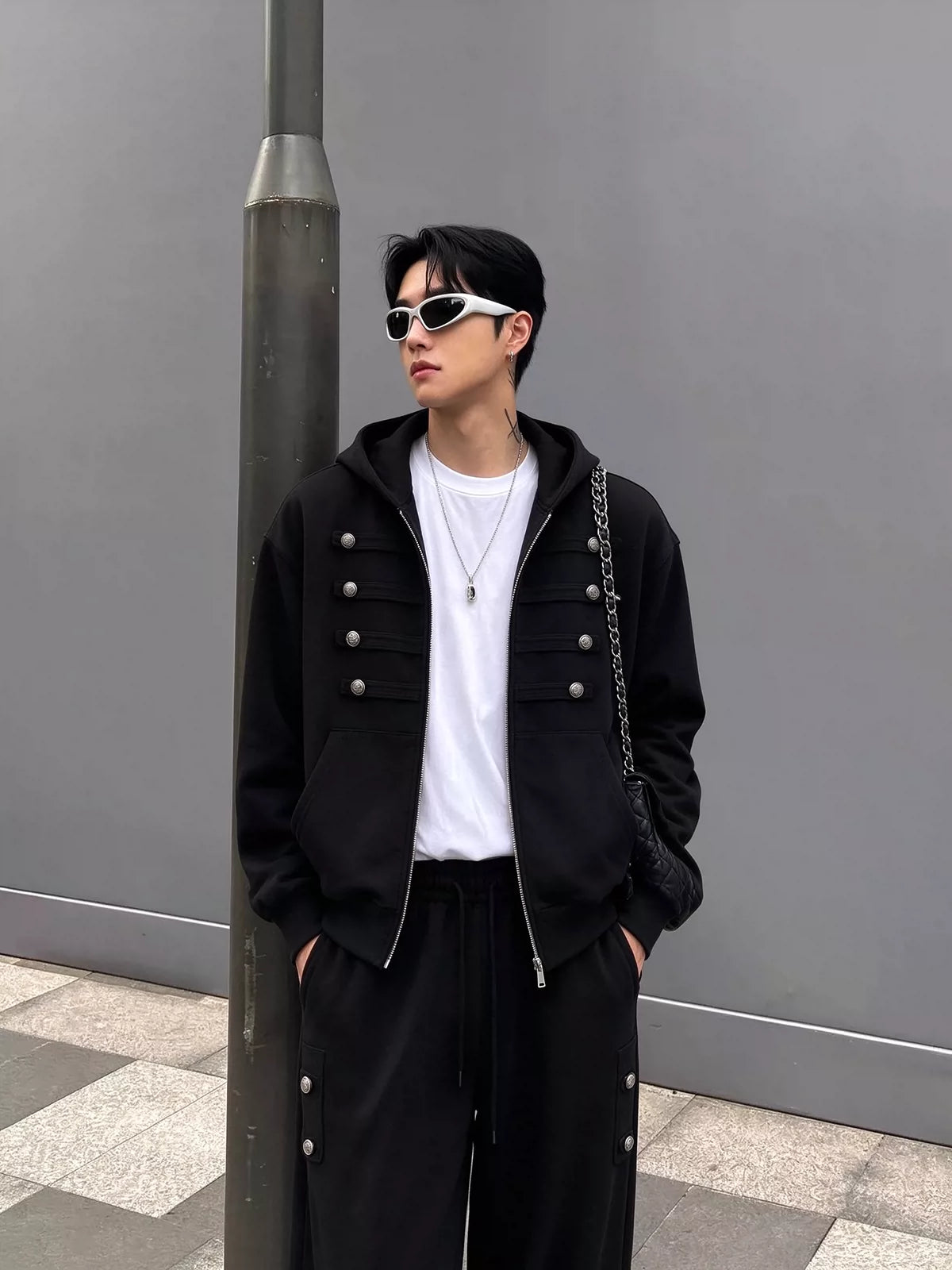 メンズの黒のジップパーカーを前開きで羽織り、白Tシャツを見せた正面の着用写真。パンツの脇ポケットにも銀ボタンが並ぶ。