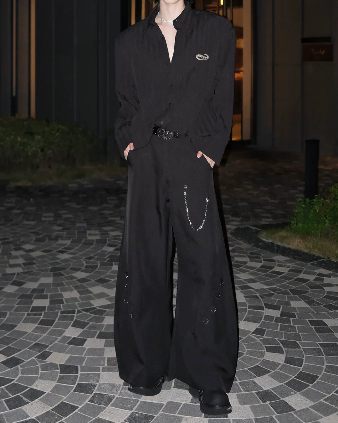 メンズの黒のシャツと黒のワイドパンツの全身コーデ。パンツは腰にチェーンと側面にリング金具、シャツは左胸に金属ロゴ付き。バックル付きベルトを合わせる。