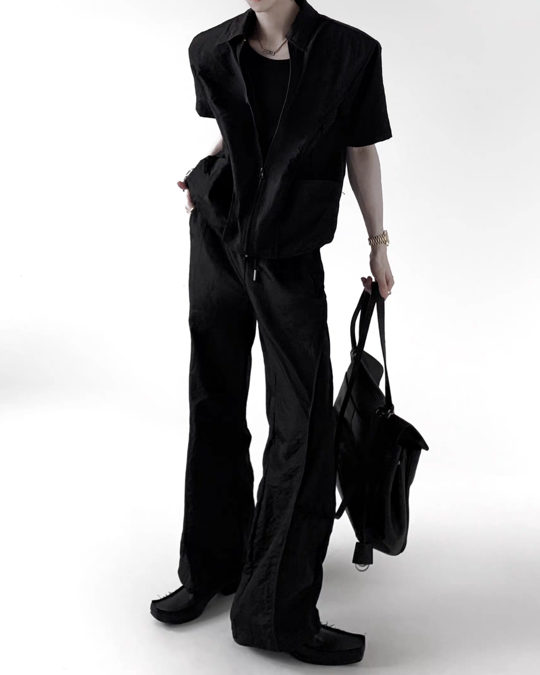 メンズの黒のジップアップ半袖シャツとワイドパンツのセットアップ。ウエストはドローコードで、黒い革のバッグを手に持っている。