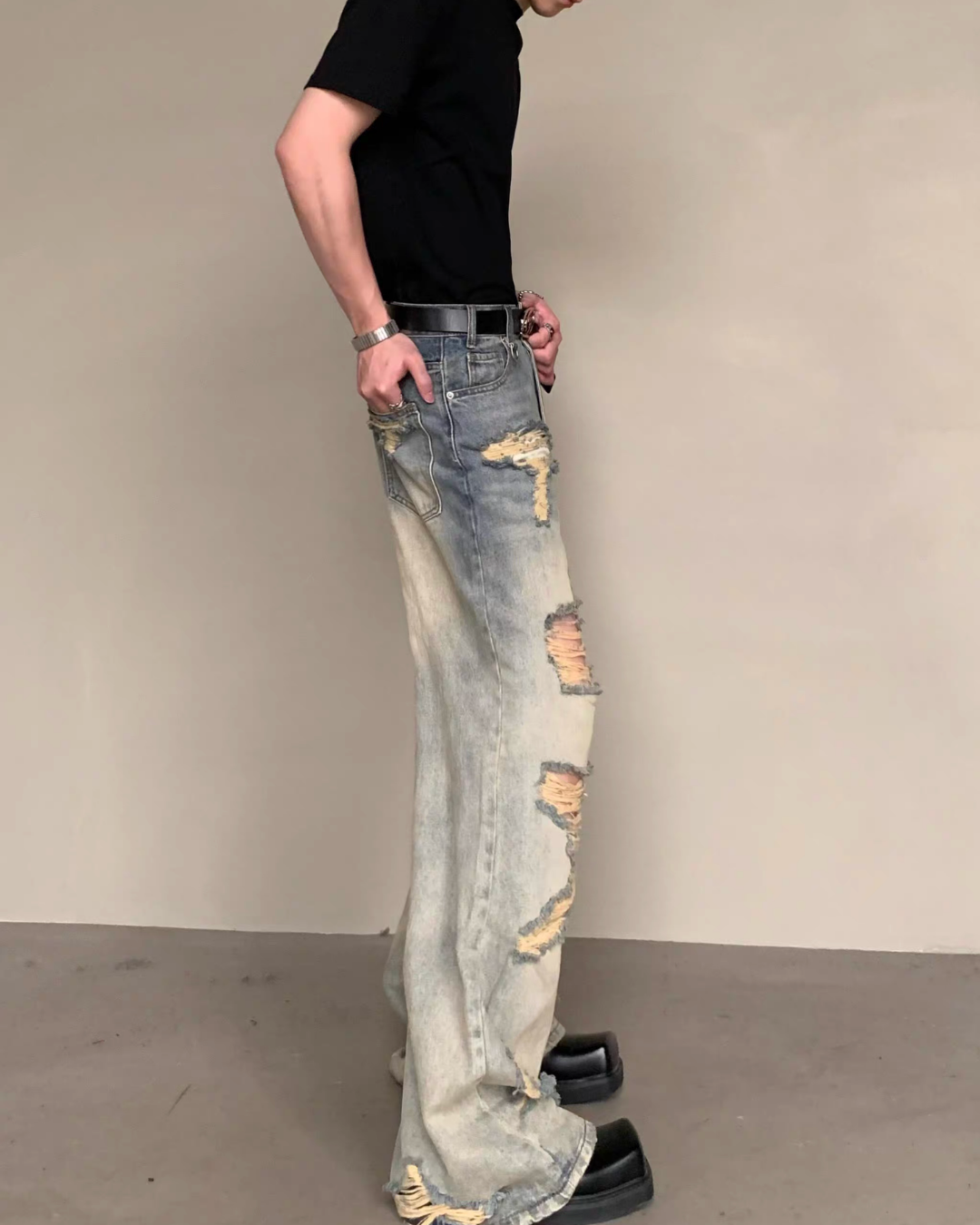 メンズの横から見たダメージデニム。ウォッシュ加工の色むらと腿・膝の裂け目、フラップ付きの後ろポケット、広がる裾が見える。