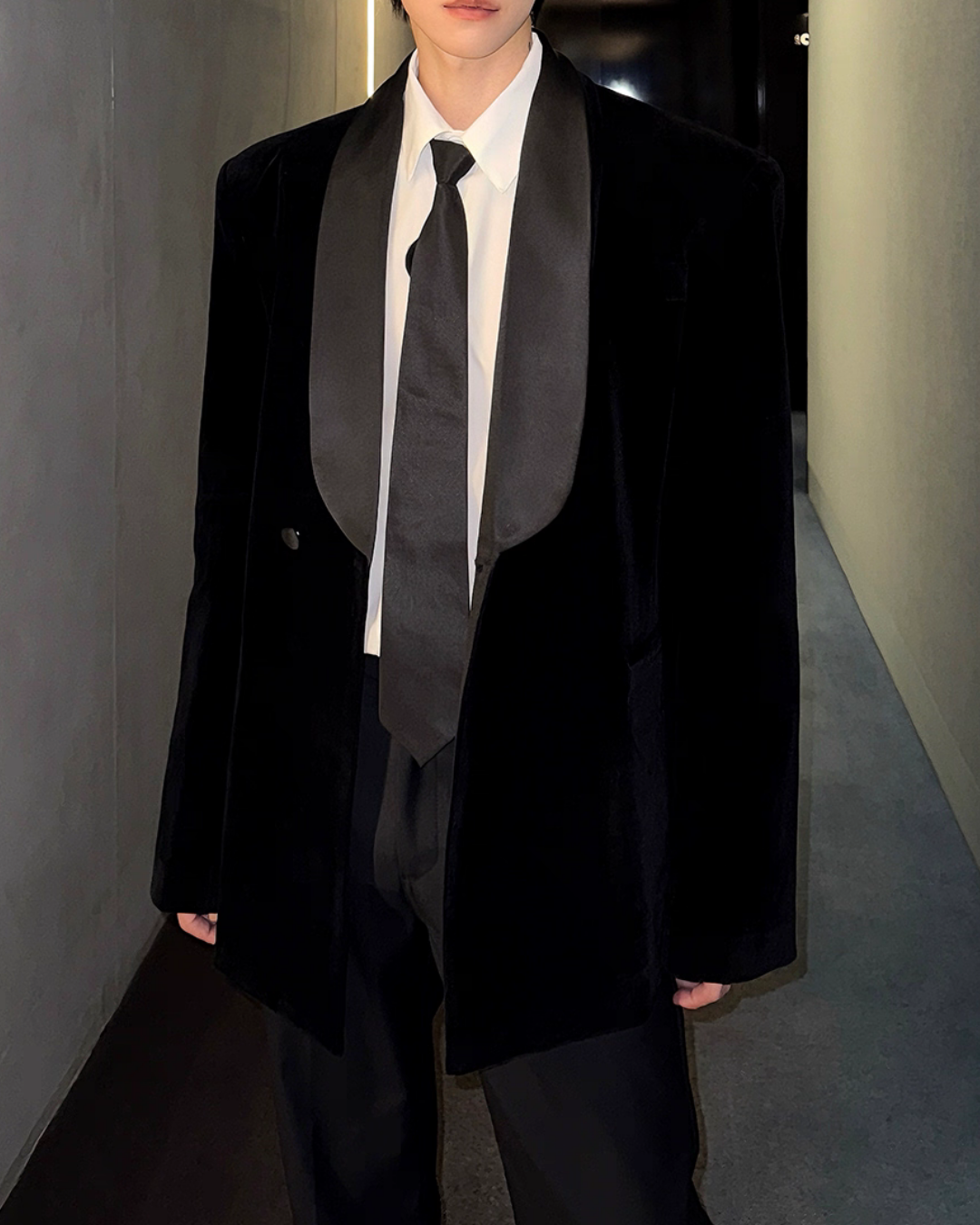 メンズのサテンのショールカラーが大きく広がる黒ベルベットのジャケット。前を開けて白シャツと黒サテンのタイを覗かせ、黒パンツを合わせる。