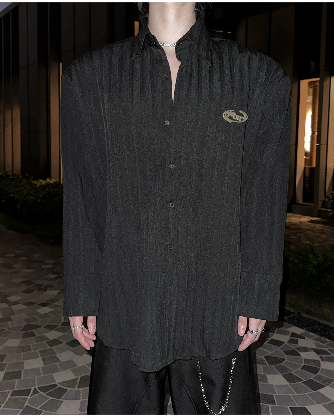 メンズの黒のシワ加工シャツの正面。ボタンを留めた前立てと尖り気味の襟、肩の張った形で、左胸に金属ロゴ。両手は下げ、黒パンツにチェーン付き。