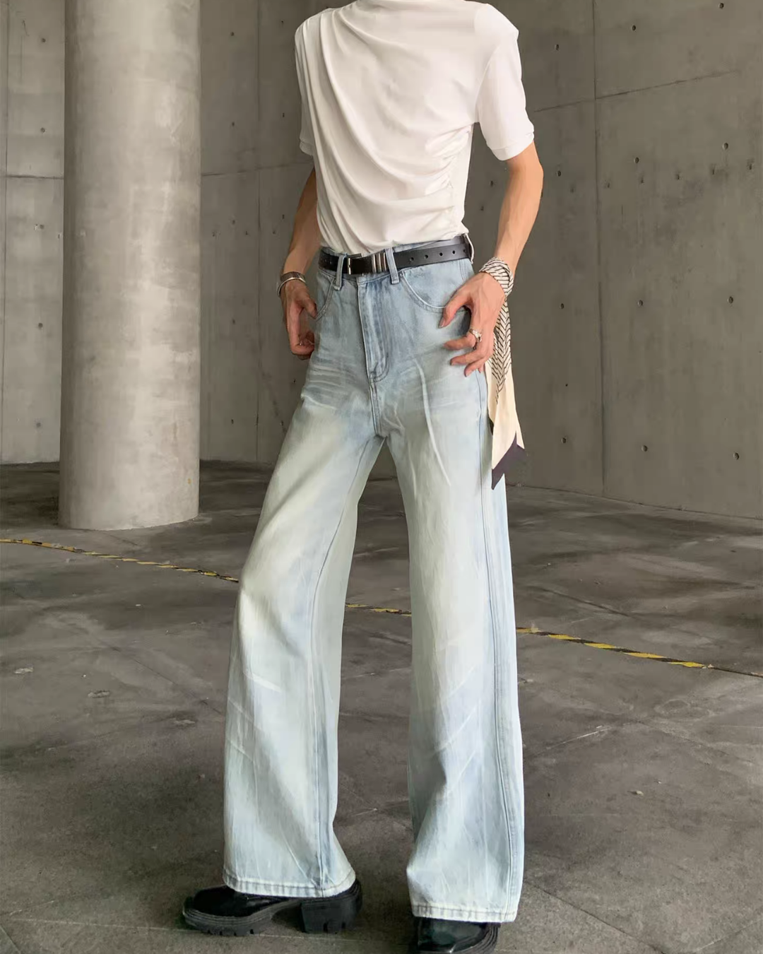 メンズの淡いブルーのデニムパンツ。両手を腰に添えた着用で、直線的なワイドシルエット。ウエストに黒ベルト、白カットソー