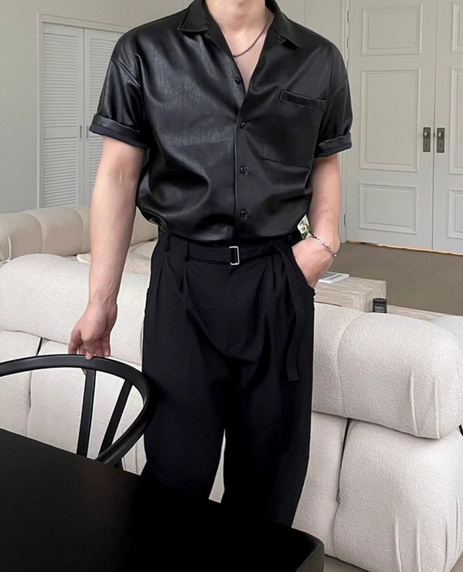 黒いレザーシャツ。半袖で袖口は折り返され、開襟で胸ポケット付き。黒いベルト付きの黒いワイドパンツとセット。