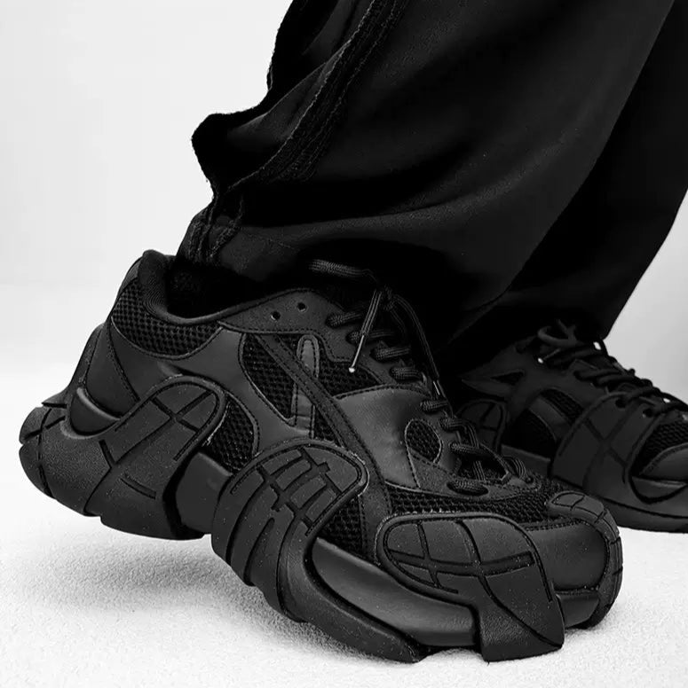 メンズの黒のスニーカーの側面斜め寄りの着用画像。メッシュとレザーの切替、細い黒の靴紐、外側に張り出すソールの立体的な造形が見える。