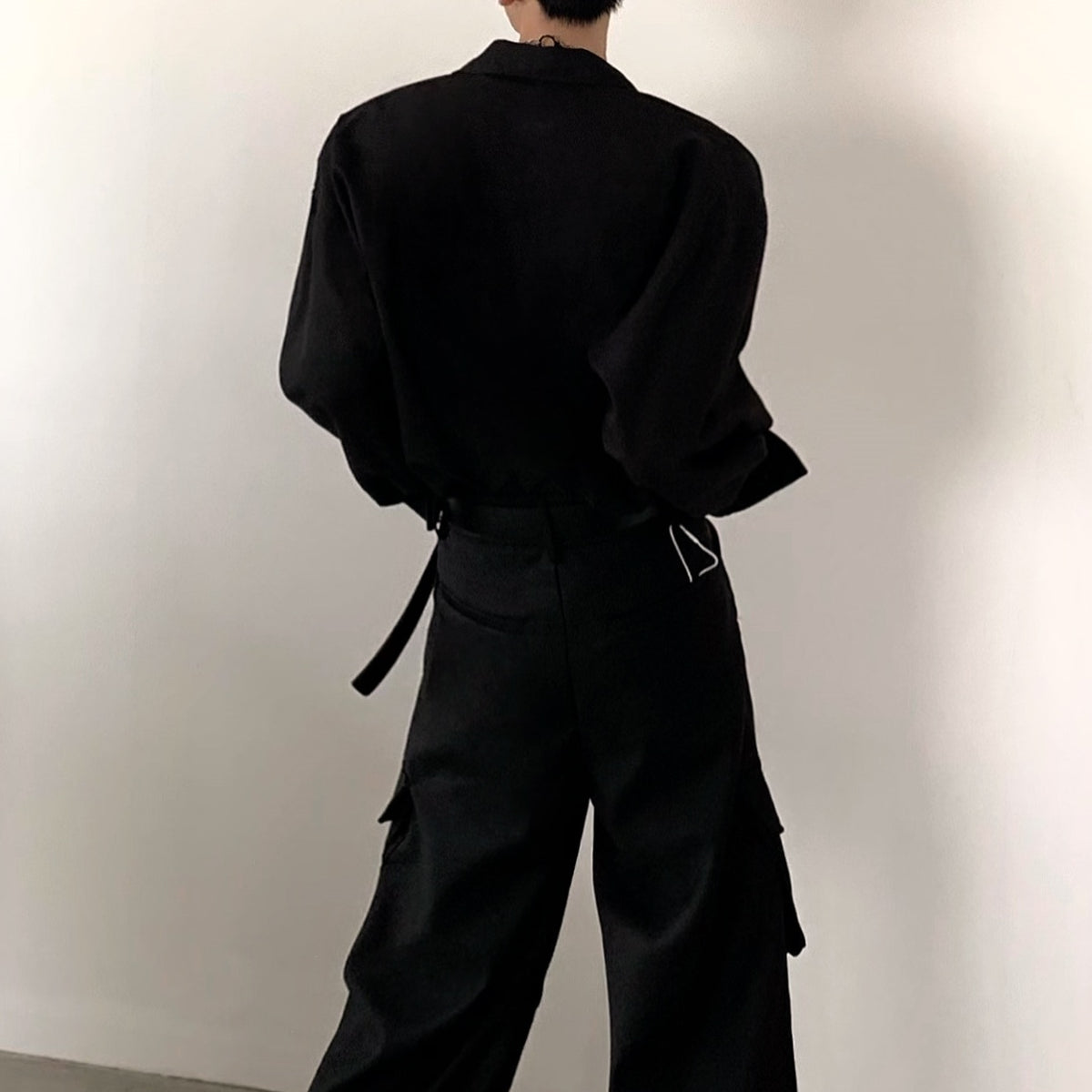 メンズの黒の長袖シャツとカーゴパンツを後ろから見たカット。肩の張った立体的なシルエットで、腰に黒ベルト、パンツ脇に大きなポケット