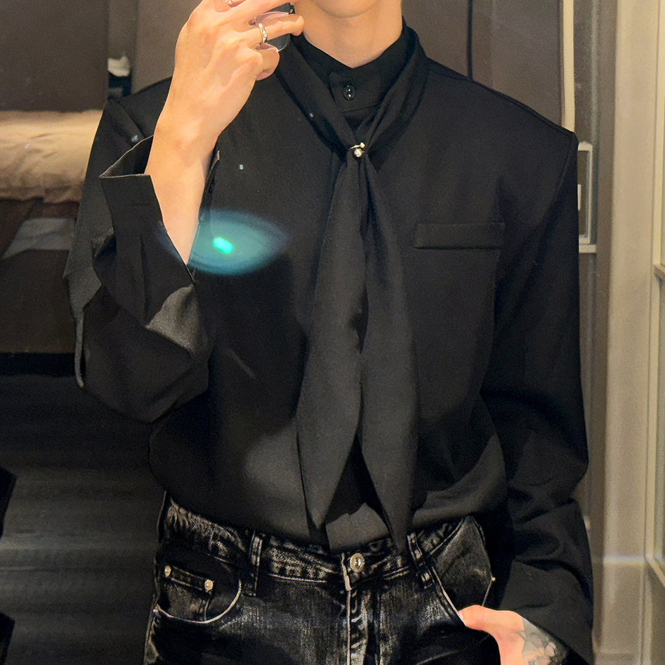 黒いオーバーサイズシャツ。Vネック、長袖で、胸元にタイデザインの装飾が施されている。前身頃にポケット付き。