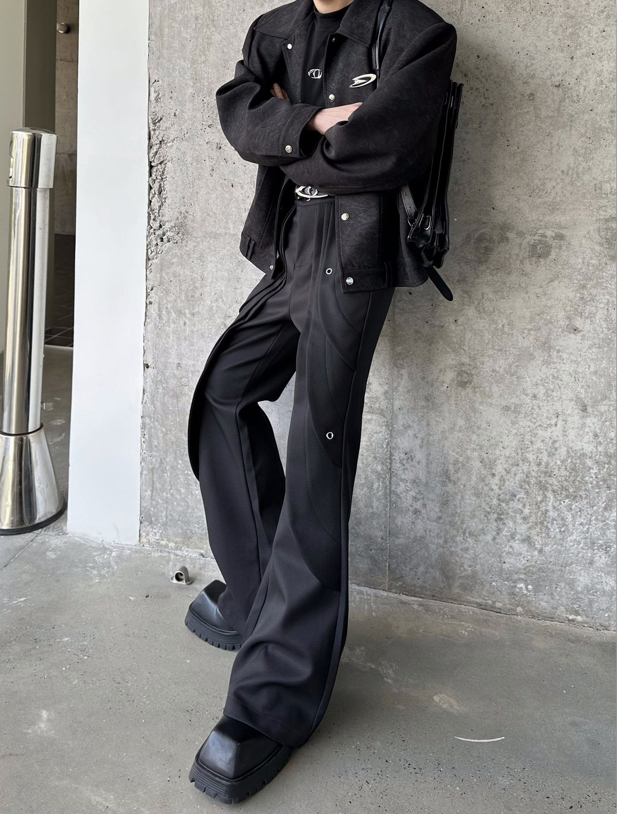 メンズの黒のフレアパンツ。腕を組んだ着用で、脇にハトメ金具と曲線の切り替えが入り、ジャケットを合わせた。