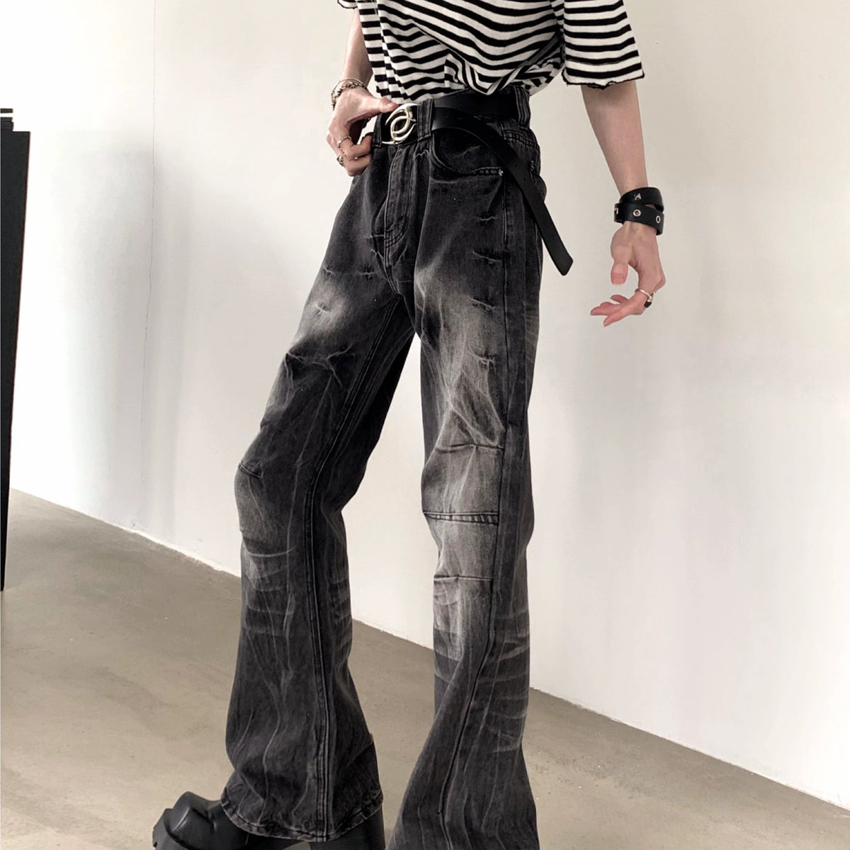 メンズの壁際で斜めに立つ着用。黒デニムのしわ加工と膝のステッチ切替が見え、黒いバングルとボーダーTシャツを合わせる。