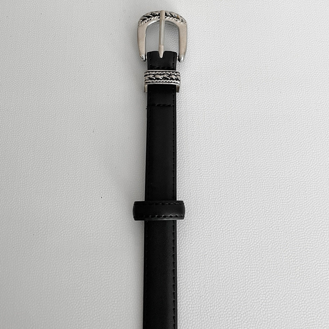 メンズの黒レザーベルトのバックル側を縦に置いた寄り画像。編み込み柄のシルバーバックルと黒のベルト通しが見える。