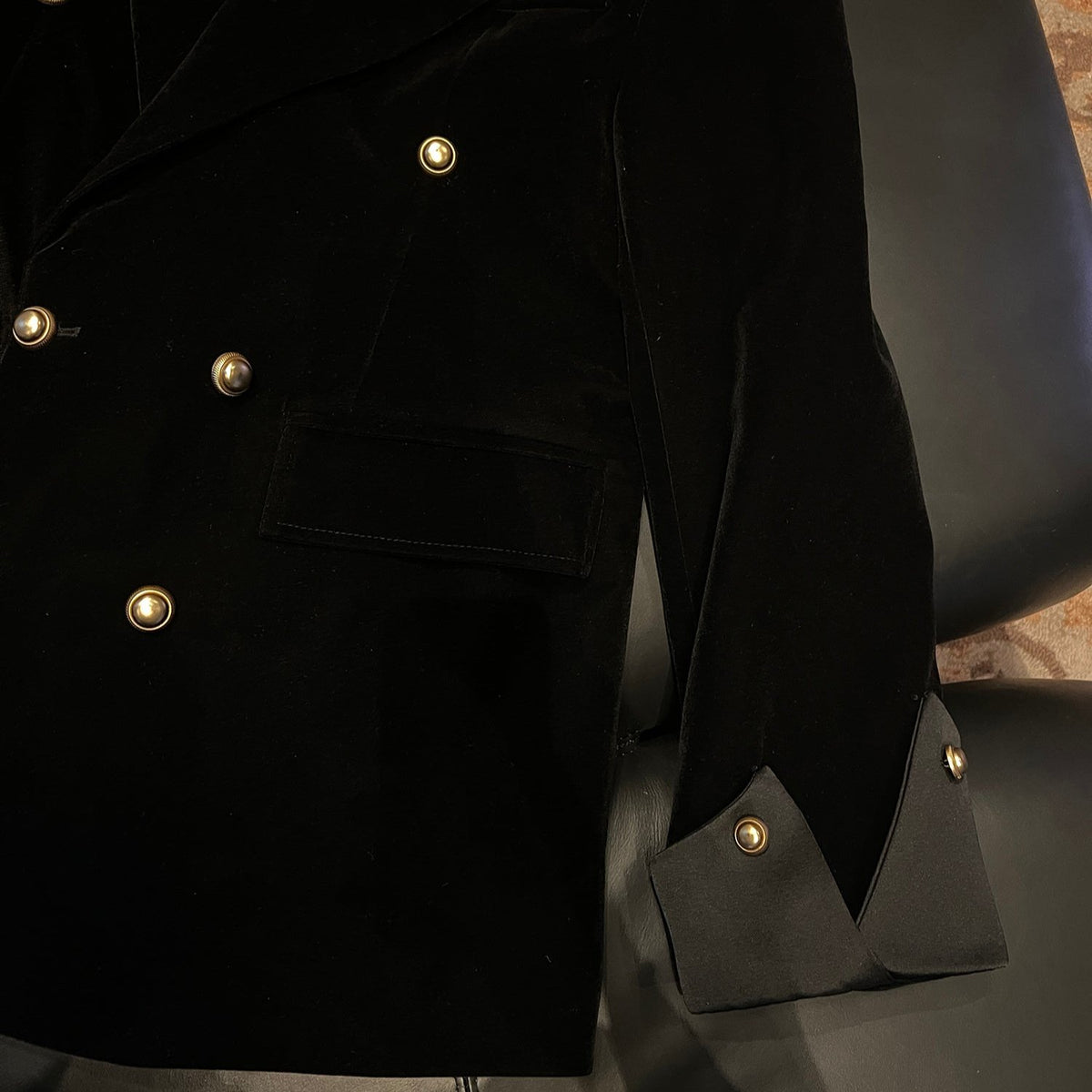 メンズの黒のジャケットを平置きにした裾と袖のアップ。金ボタン、フラップポケット、袖口の金ボタンが付いた三角形のサテン調カフスを寄りで見せる。