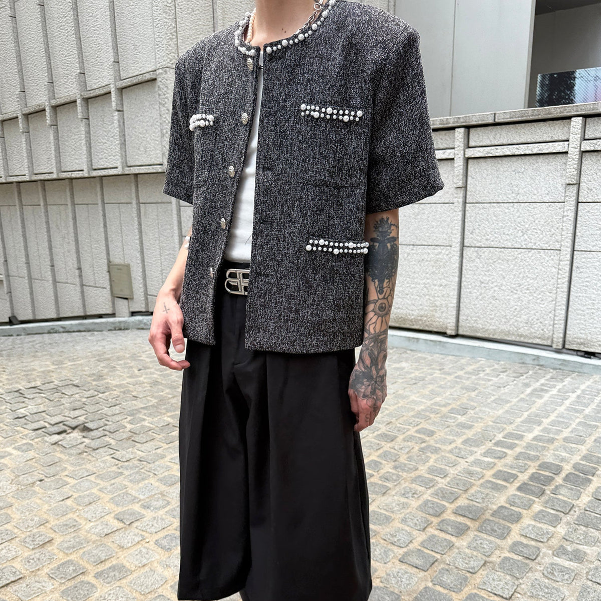 メンズの白いトップスに重ねた黒ツイードの半袖ジャケット。襟ぐりとポケットのパール、タックの入った黒パンツ