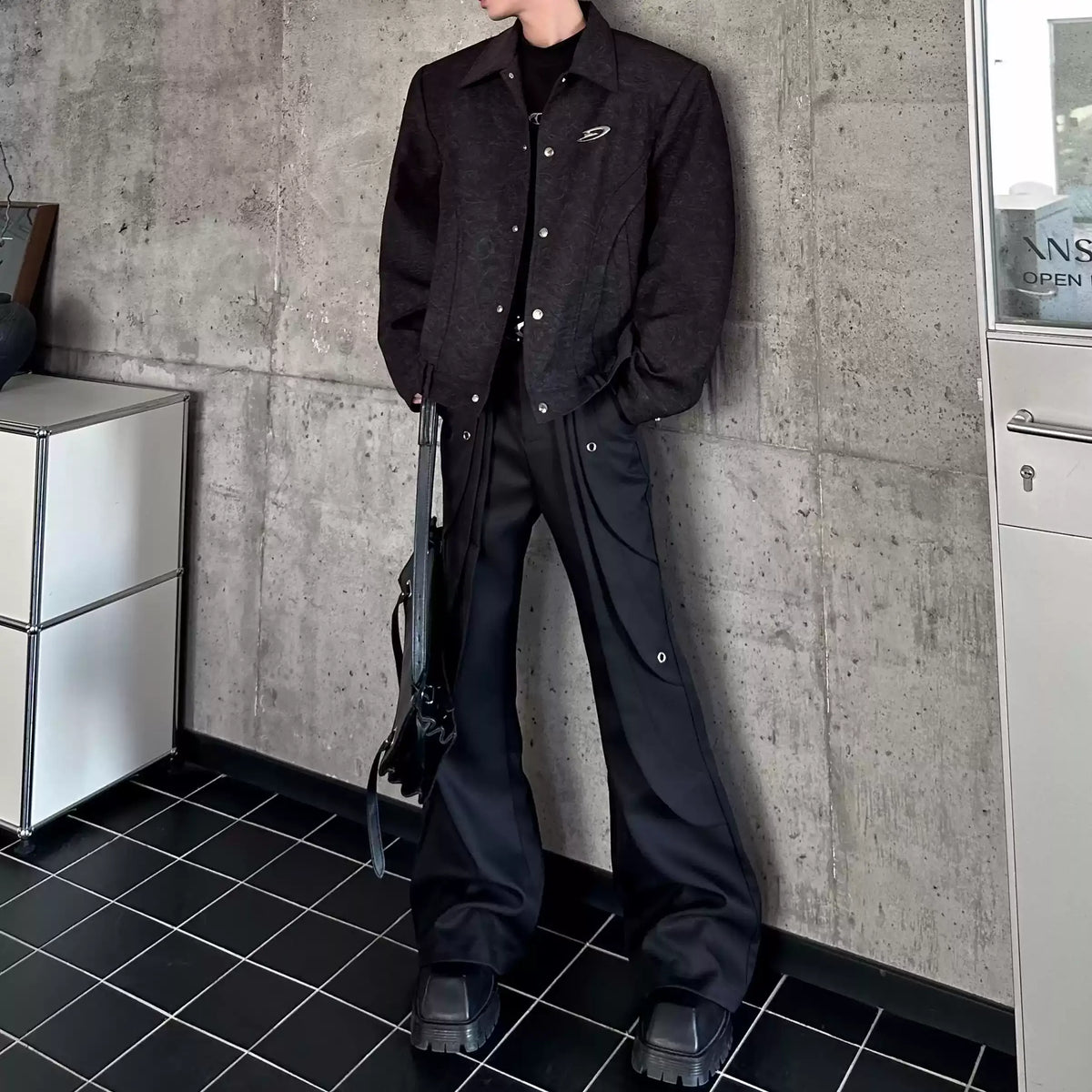 メンズの黒のフレアパンツ。脇にハトメ金具と曲線の切り替えが入り、黒の織り柄ジャケットとバッグを合わせた全身。