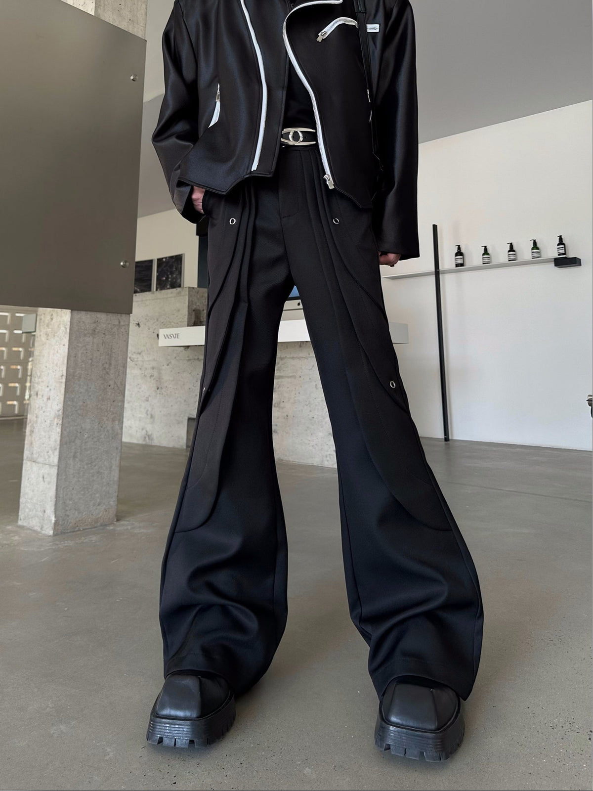 メンズの黒のワイドなフレアパンツ。ハトメ金具と曲線の切り替えが入り、白いジップ付きの黒ジャケットを合わせた寄り。