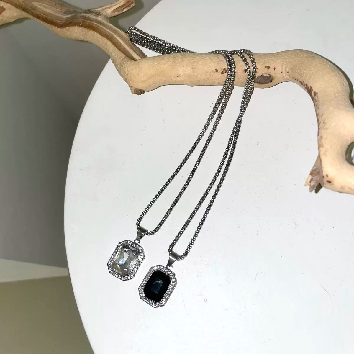 メンズの流木に掛けた、透明石と黒石のペンダント付きシルバーネックレス2本。白い円形の台の上に並べた全体写真。