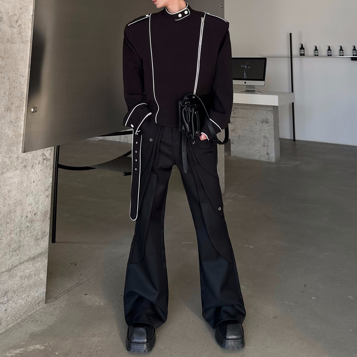メンズの黒のフレアパンツ。腰にハトメ金具付きのレイヤーがあり、白パイピングの黒トップスと黒い小型バッグを合わせた全身。