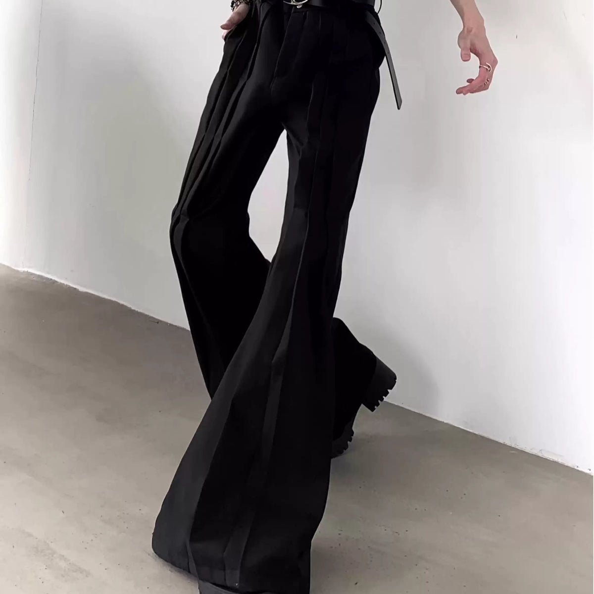 メンズの歩く姿勢で写した黒フレアスラックス。縦に走る複数のプリーツが入り、裾へ向かって大きく広がる。