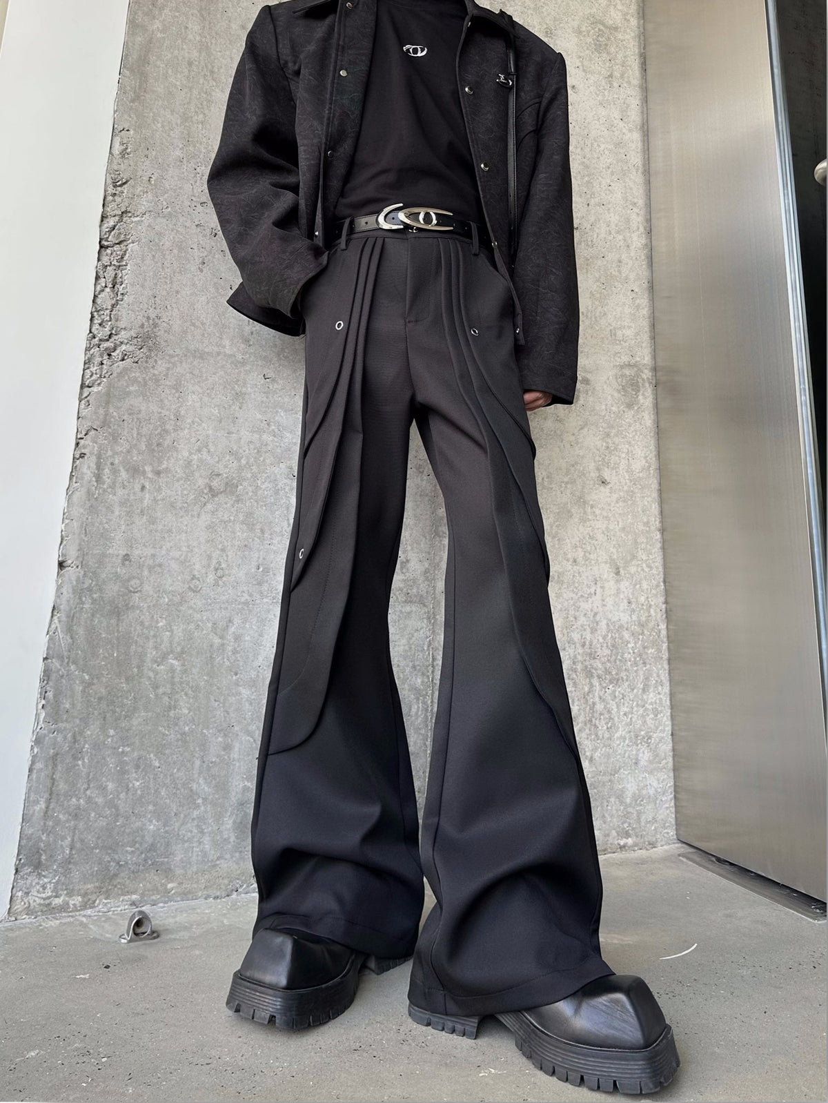 メンズの黒のフレアパンツの正面の寄り。腰に重なるプリーツとハトメ金具があり、銀色バックルのベルトを合わせた。