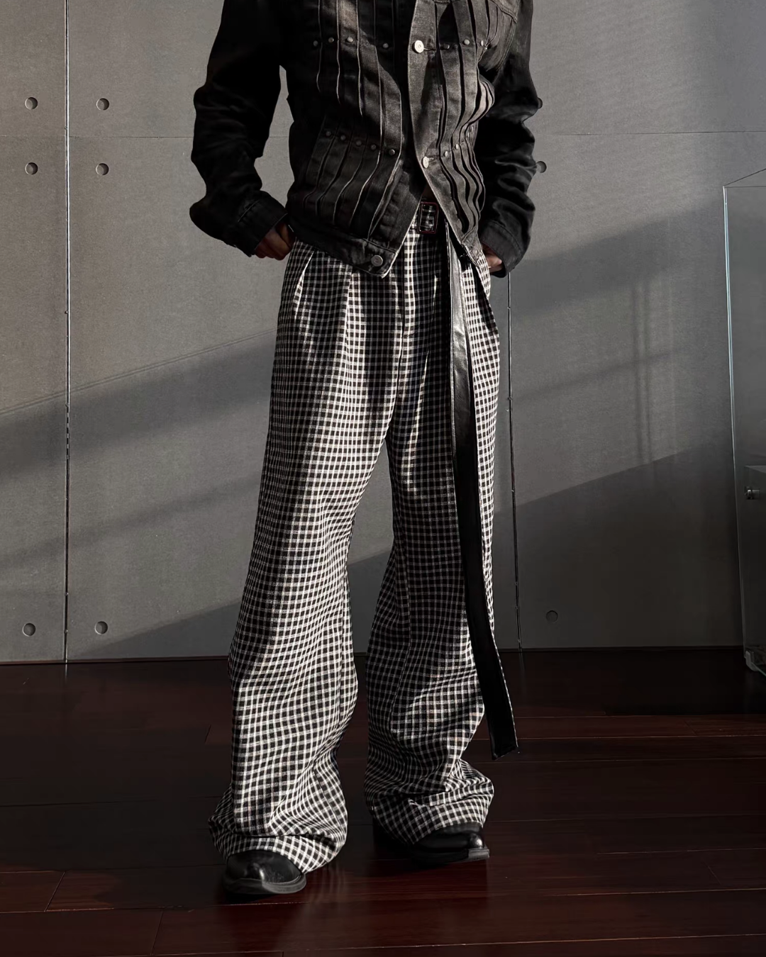 メンズの白黒チェックのワイドパンツ。黒革のベルトが腰から垂れ、タックの入った腰から裾まで広がる。黒デニムジャケットと黒の革靴。