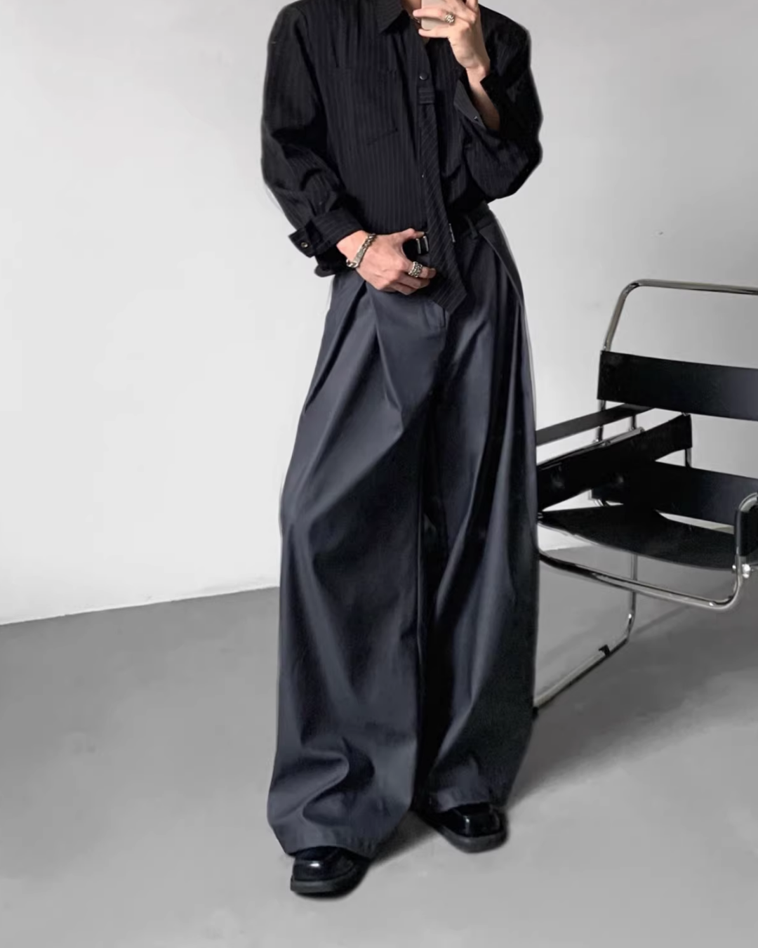 メンズの黒のピンストライプシャツに濃いグレーのワイドスラックスを合わせた全身着用画像。スラックスは腰から裾にかけて大きなサイドプリーツが入り、ドレープが特徴的。