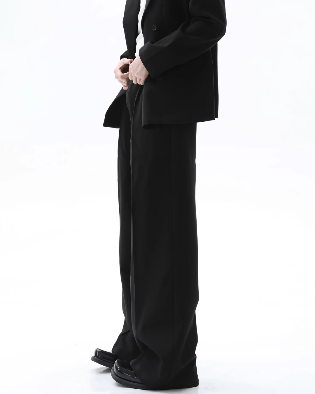 メンズの黒のワイドパンツを横から捉えた下半身。ウエストにタック、裾は床に届く長さ。丈短めのジャケットと角のある黒シューズ。