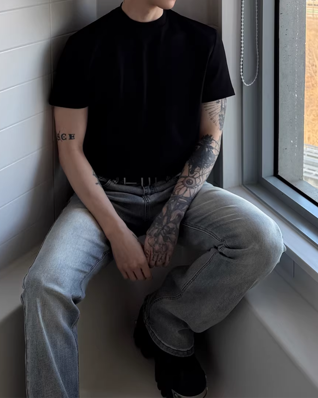 黒の半袖クルーネックTシャツ。直筒シルエットでコットン素材。ウィンドウ際での着用写真。