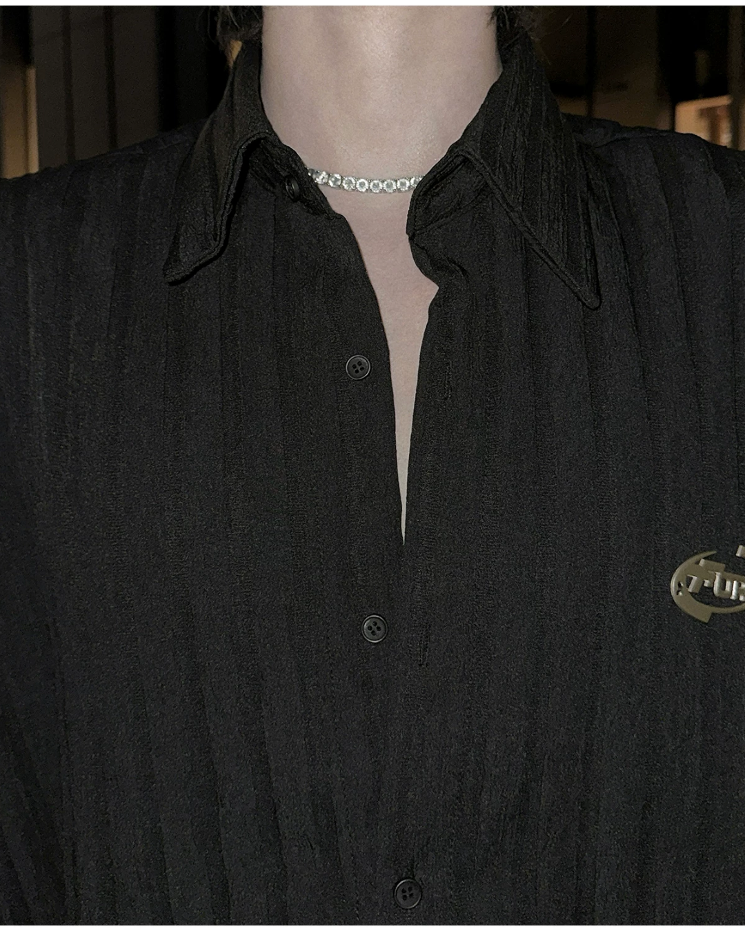 メンズの襟元を正面から寄りで写した黒シャツ。左右に先の尖った襟と黒ボタン、縦のプリーツ調の生地、右胸の金属ロゴが見える。