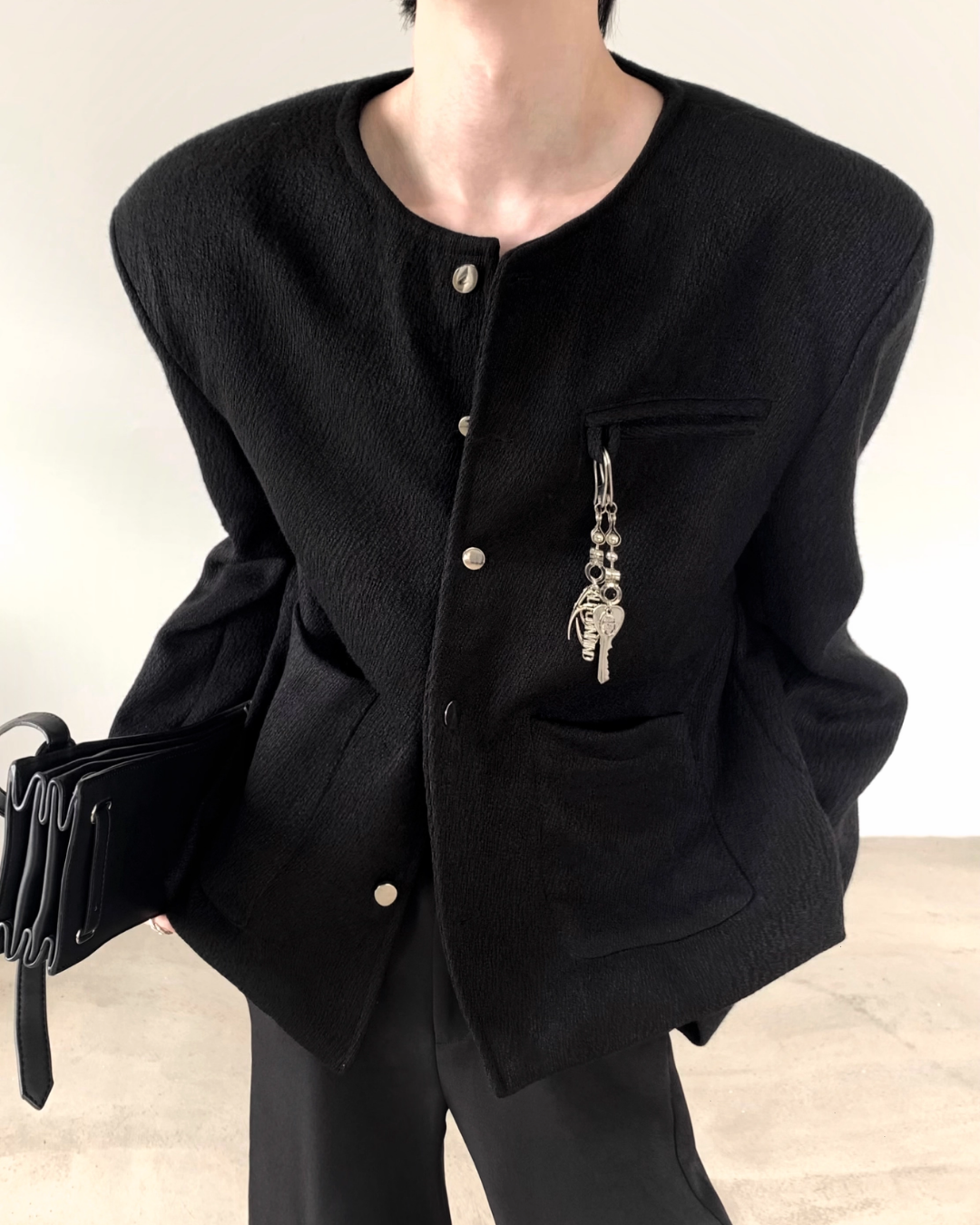 黒のノーカラージャケット。前立てにシルバー金具ボタン、胸ポケット付き。シルバーチェーンブローチが装着されている。