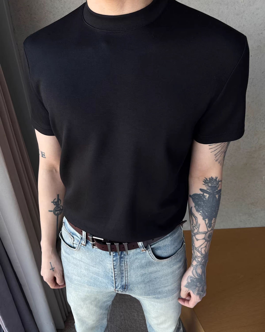 黒のクルーネック半袖カットソー。シンプルで直線的なシルエット。着用者は淡色デニムとブラウンレザーベルトを合わせている