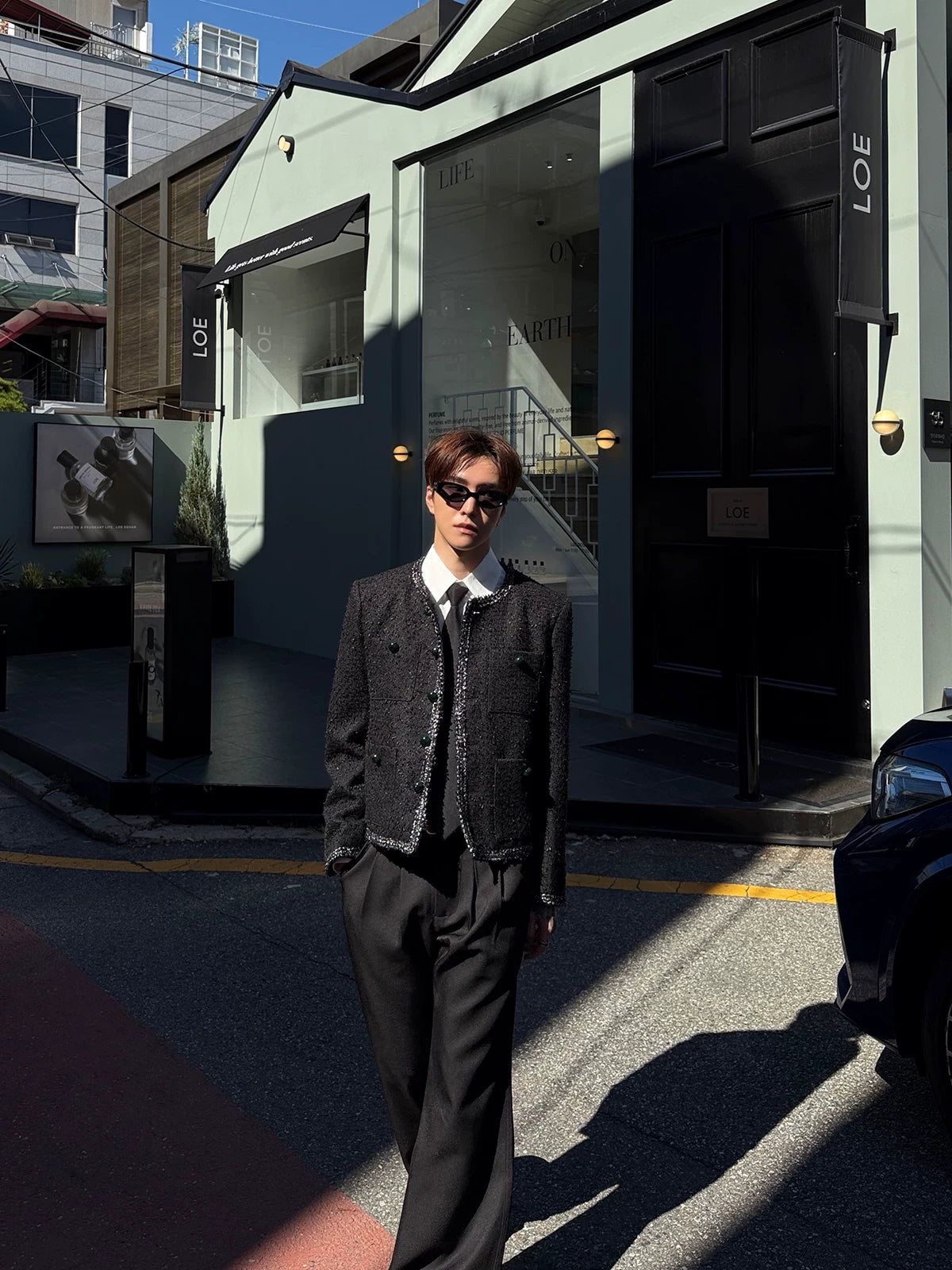 メンズの黒いツイード調ジャケットと黒のワイドパンツ。白シャツとネクタイを合わせ、店の前で正面に立つ全身。