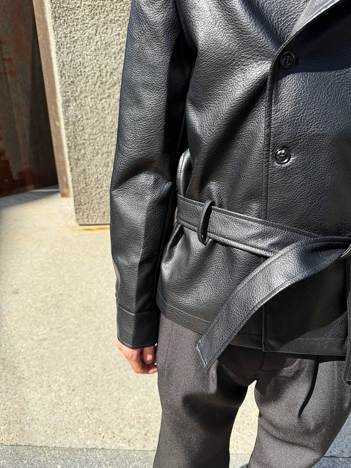 メンズの黒いレザー調ジャケットの腰まわりの寄り。シボのある革の表面、ベルト通しと結んだベルト、袖口の折り返しを横から写す。