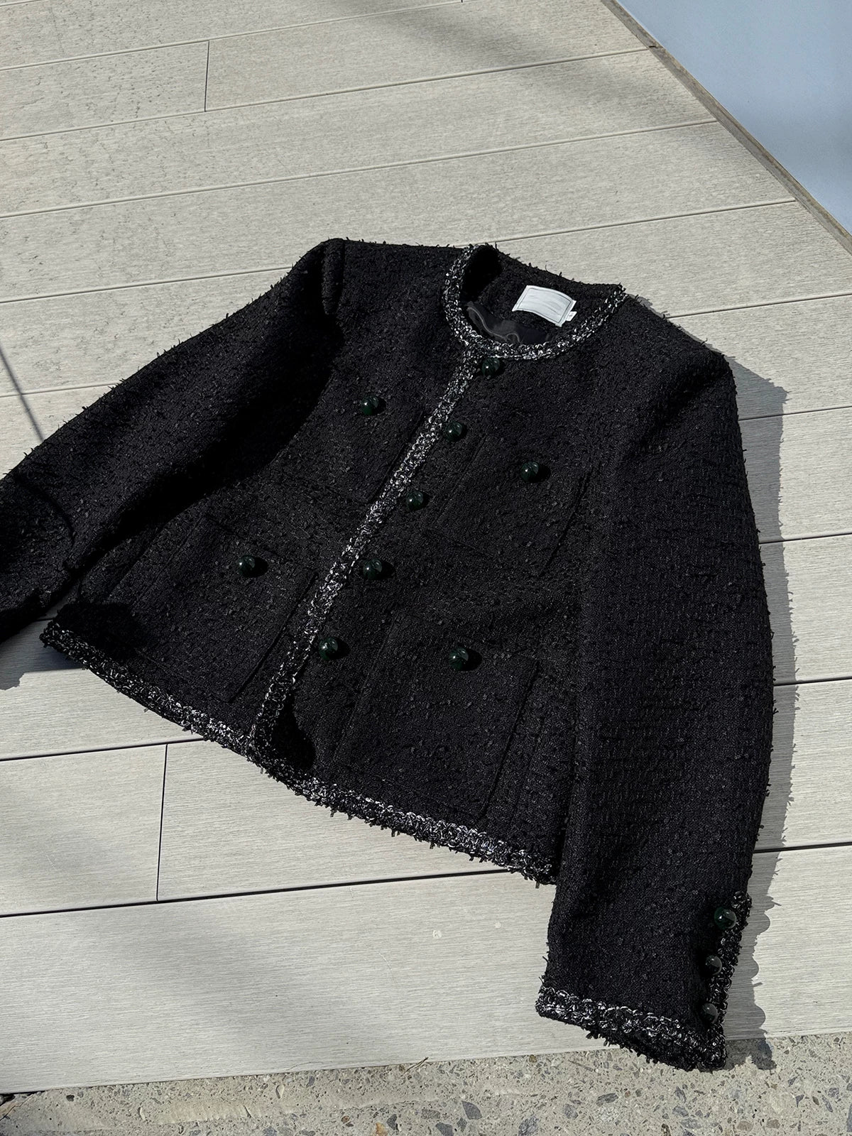 メンズの黒いツイード調ノーカラージャケットの平置き。前に緑のボタンが並び、胸と腰に4つのポケット、袖口にもボタンが付く。