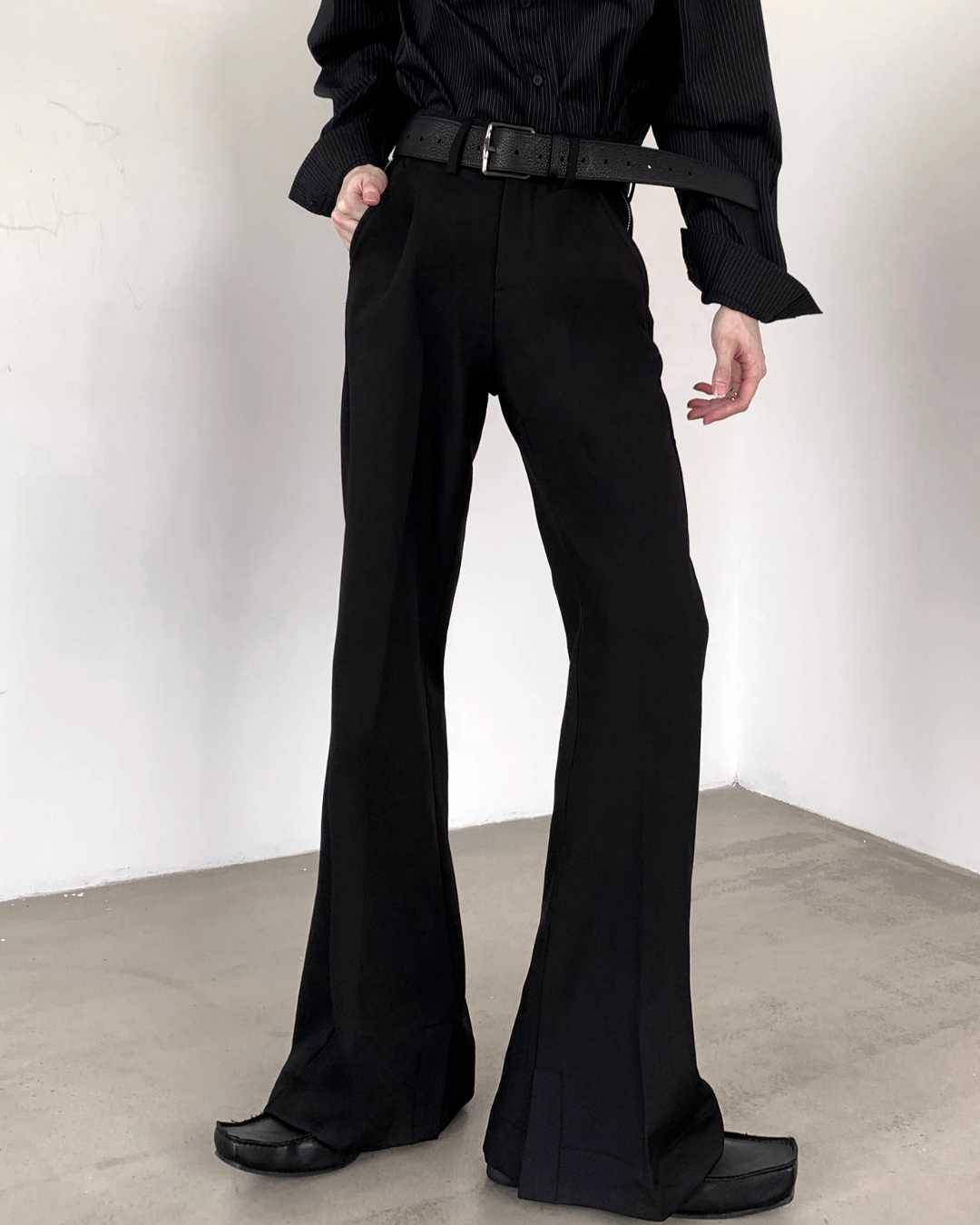 メンズの黒のハイウエストのフレアパンツ。銀の尾錠の黒ベルトを締め、タック入りで、裾のスリットが見える正面からの着用写真