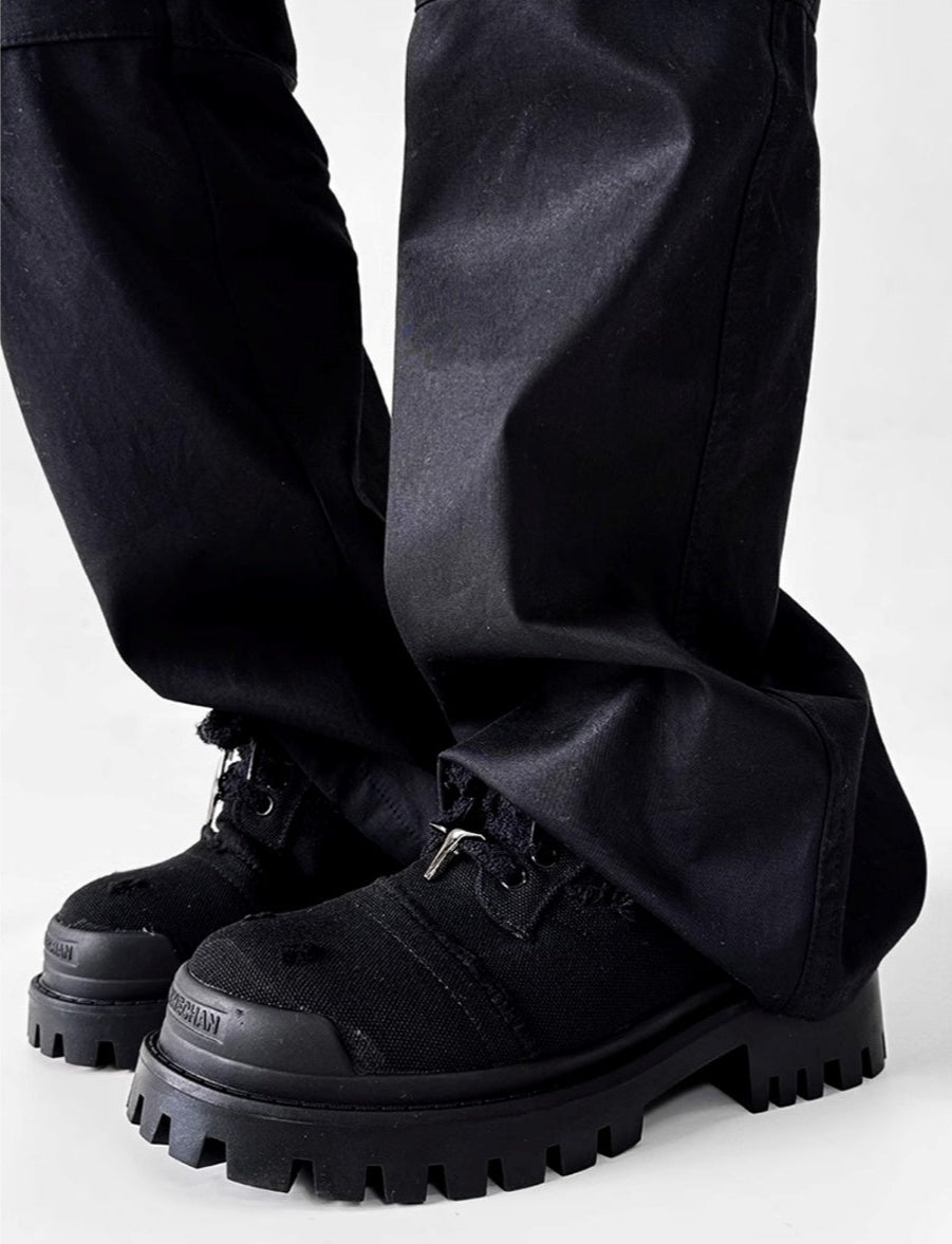 メンズの黒パンツの裾から出た黒の厚底スニーカーを横から写した着用画像。ほつれ加工の靴紐、星形の銀色金具、ラグソールが見える。