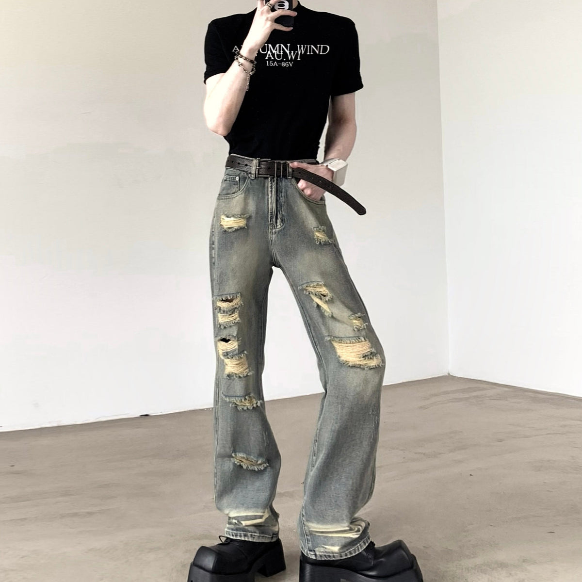 黒のロゴプリントクルーネックTシャツ。半袖で胸部に白抜き文字のデザイン。グレーのダメージデニムとベルト合わせのスタイリング。
