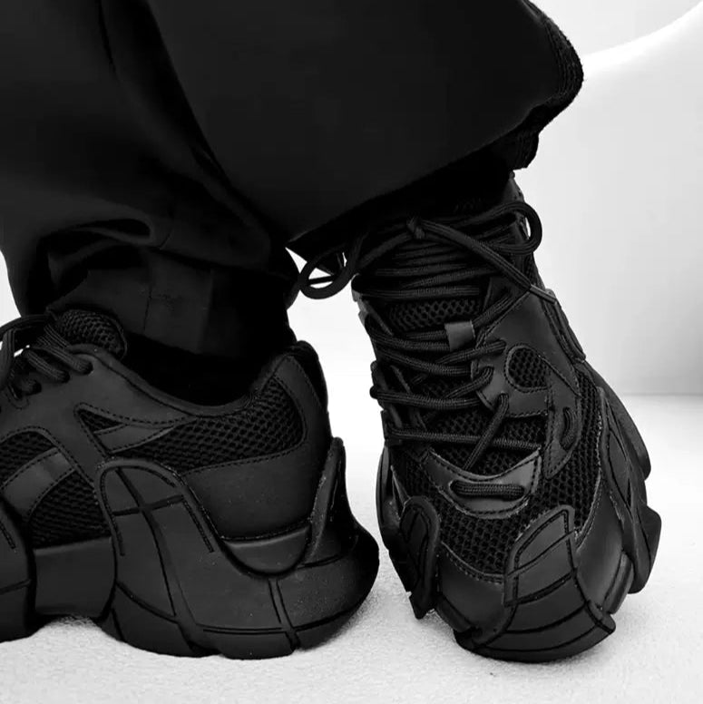 メンズの黒のスニーカーを両足で履き、片足のかかとと側面を見せた画像。黒のメッシュとレザーに、段のあるソール装飾が重なる。