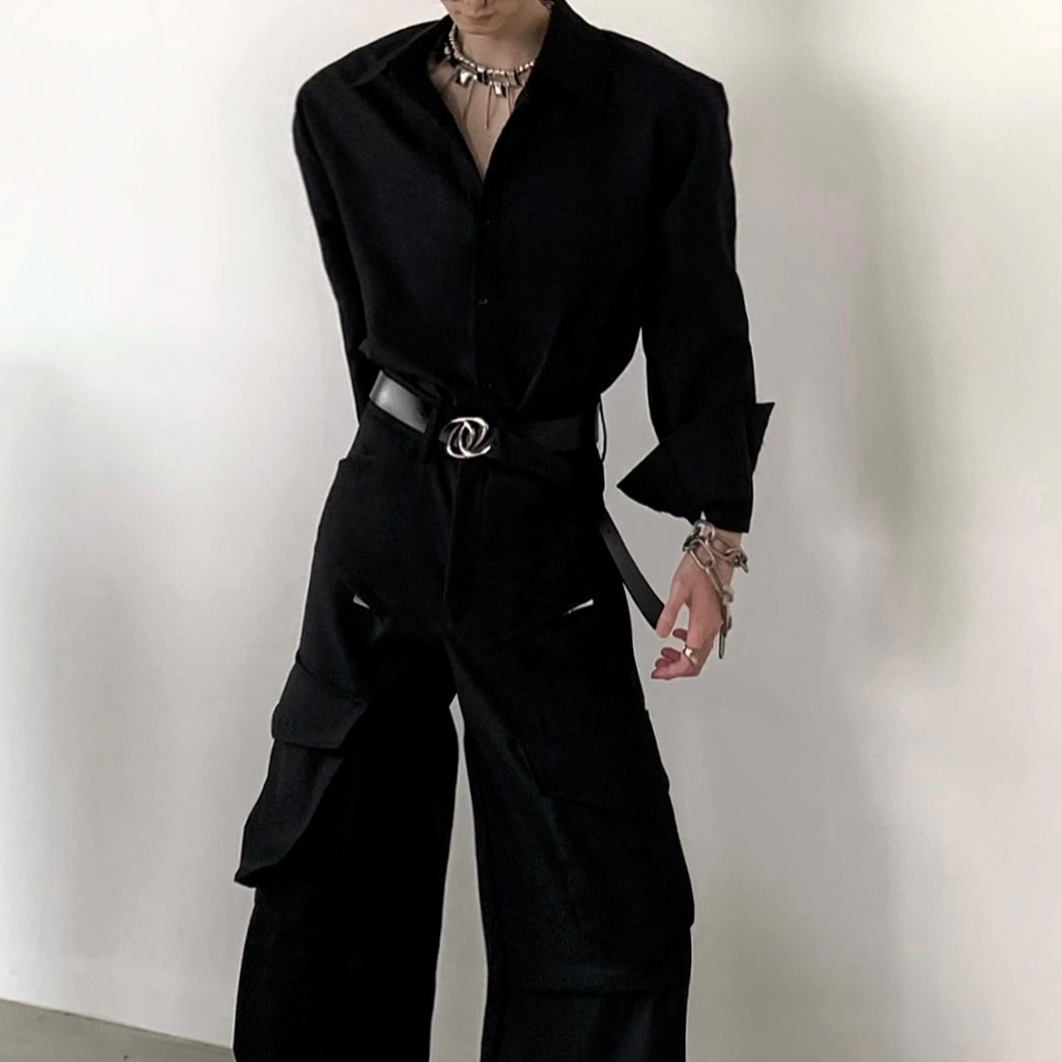 メンズの黒の長袖シャツとワイドなカーゴパンツの全身寄りカット。肩の張った形、折り返したカフス、シルバーバックルの黒ベルト