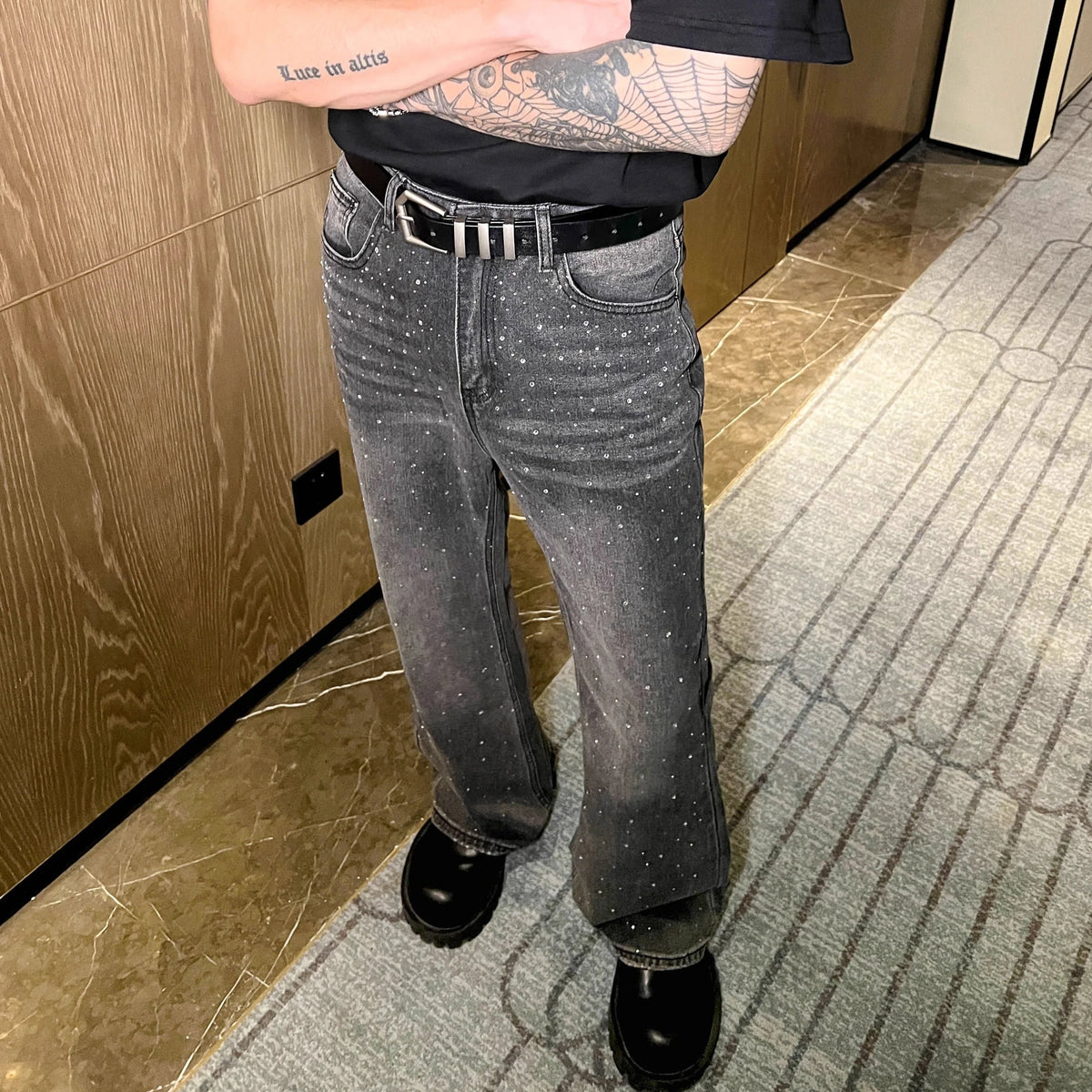 メンズのグレーのデニムパンツ。全体にクリスタルストーン装飾が施され、ストレートシルエット。黒ベルト合わせた着用写真。