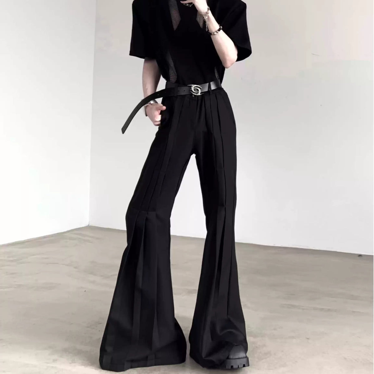 メンズの黒の半袖トップスと、縦のプリーツが入った黒のフレアスラックスの全身。シルバーバックルの黒ベルトを締めている。