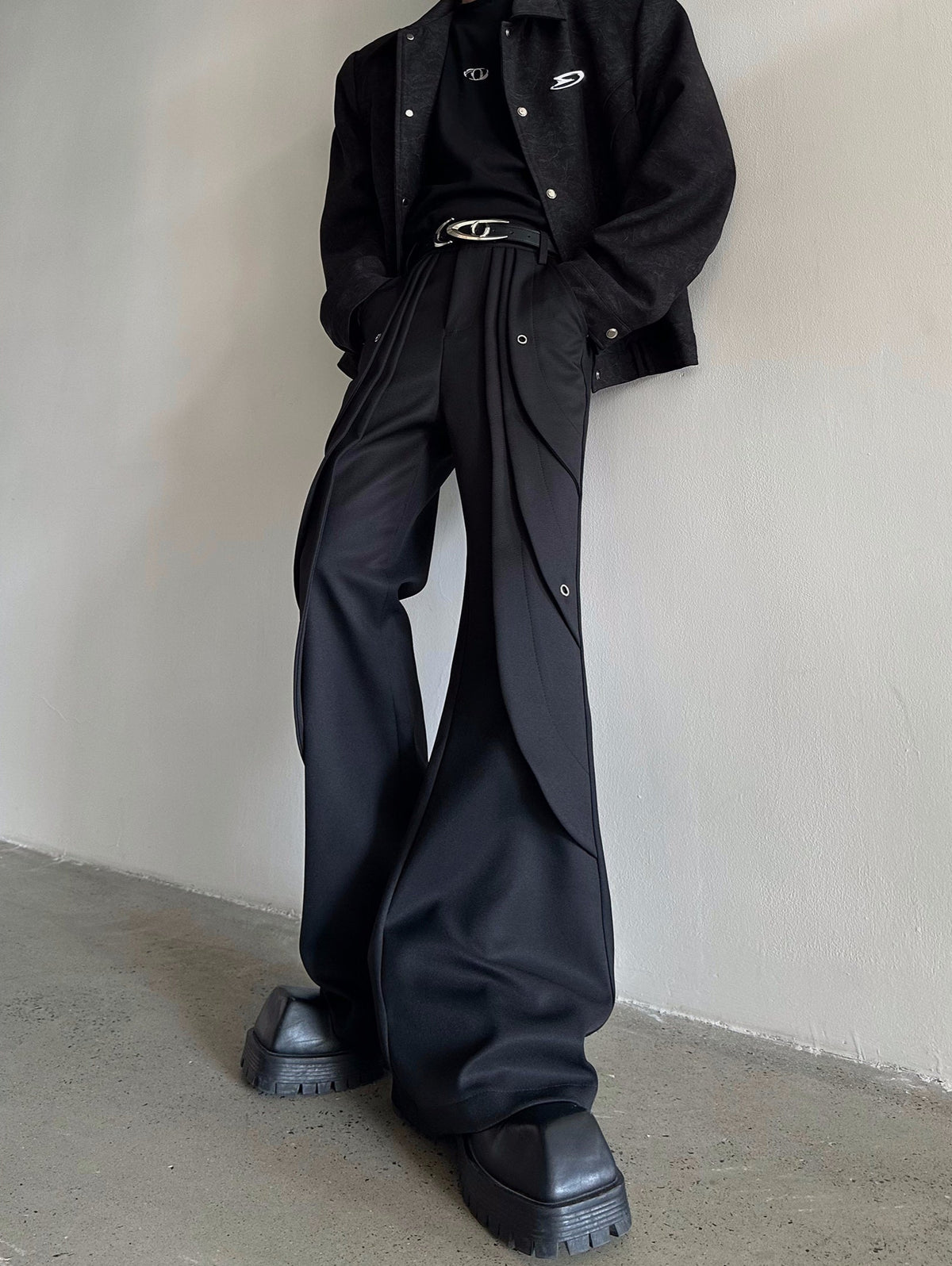 メンズの黒のフレアパンツの着用。腰にプリーツ状のレイヤーとハトメ金具があり、黒の織り柄ジャケットとベルトを合わせた。