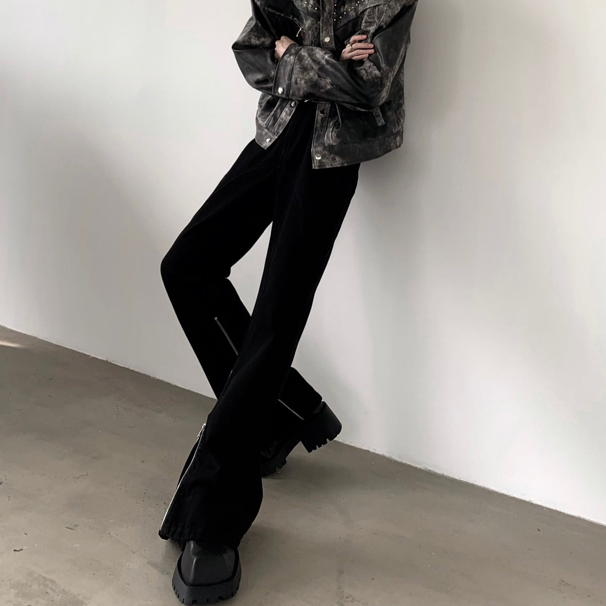黒のフレアパンツを壁にもたれて脚を交差した着用。すその脇にファスナーがあり、ダメージ加工風のジャケットを合わせる。