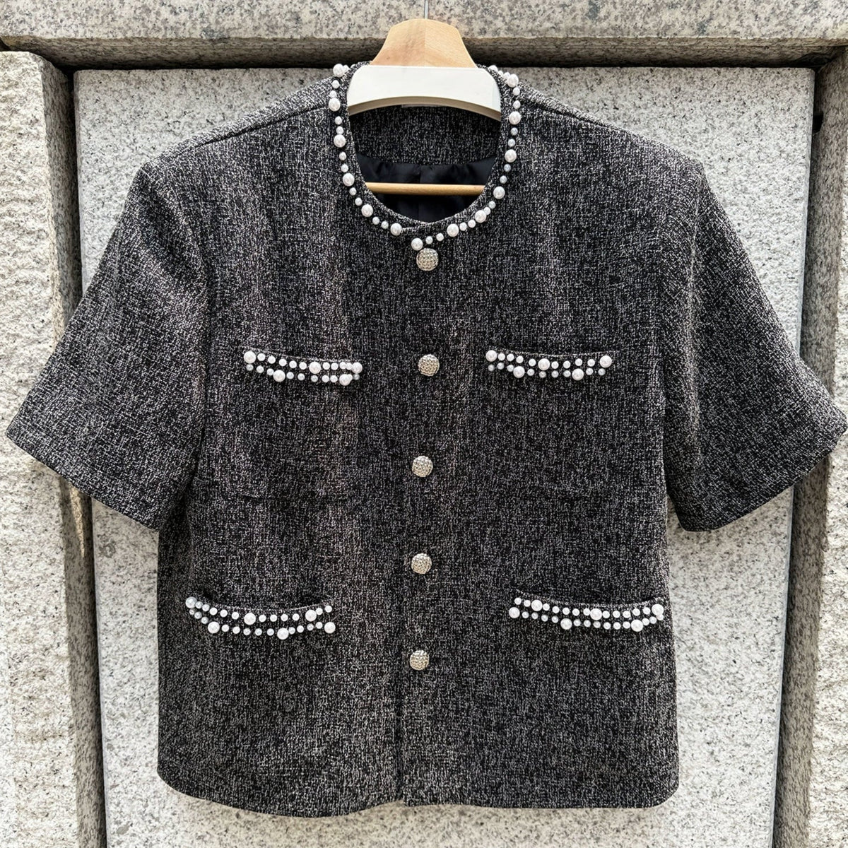メンズのハンガーに掛けた黒ツイードの半袖ジャケット。襟ぐりと4つのポケット口にパール、前立てに銀のボタン5つ