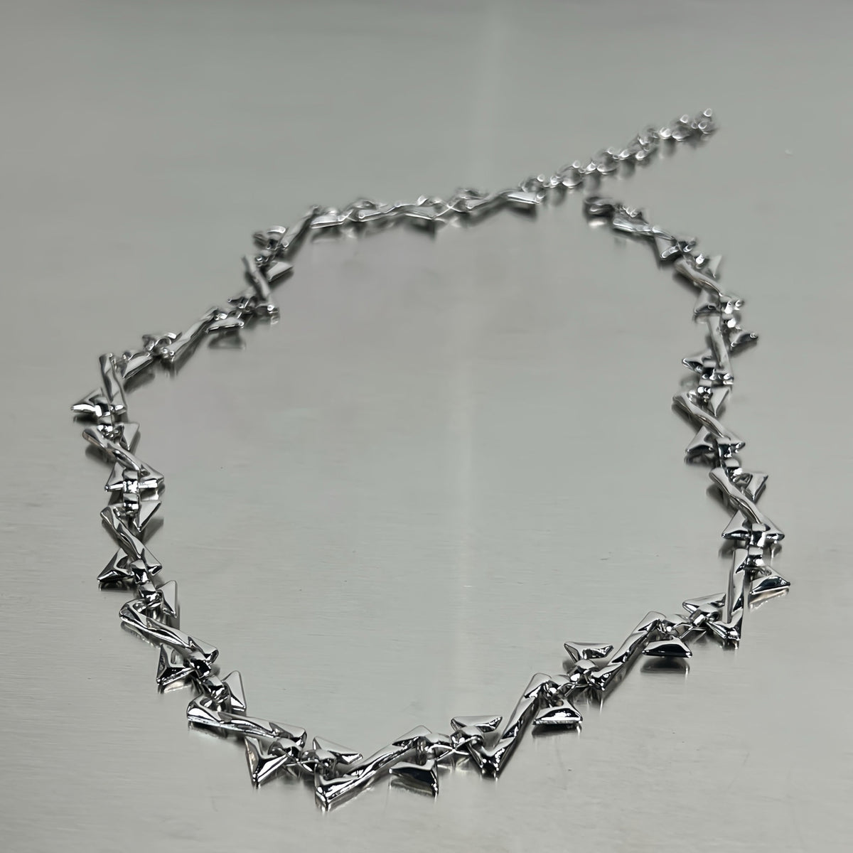 メンズのシルバーのネックレスを金属板の上に置いた平置きカット。三角形のパーツが連なり、片側にアジャスターチェーン付き
