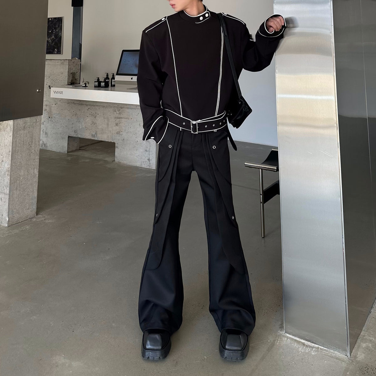 メンズの黒のフレアパンツ。腰に重なる布とハトメ金具が付き、白パイピングの黒トップスとベルトを合わせた全身。