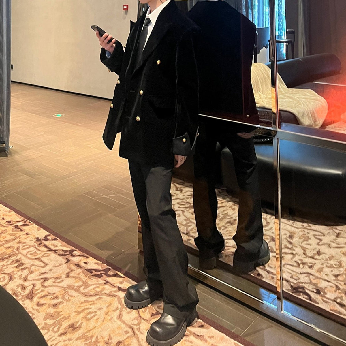 メンズの黒のダブルブレストジャケットを横向きに立って着用。ゆとりのある肩と金ボタンが見え、黒のパンツと厚底の黒い靴を合わせている。